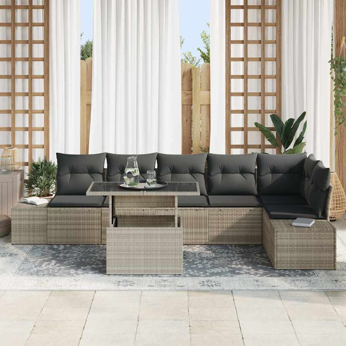 Outdoor-Rattansofa mit grauen Kissen, Glastisch mit Getränken, vor weißen Vorhängen und Holzgitter.