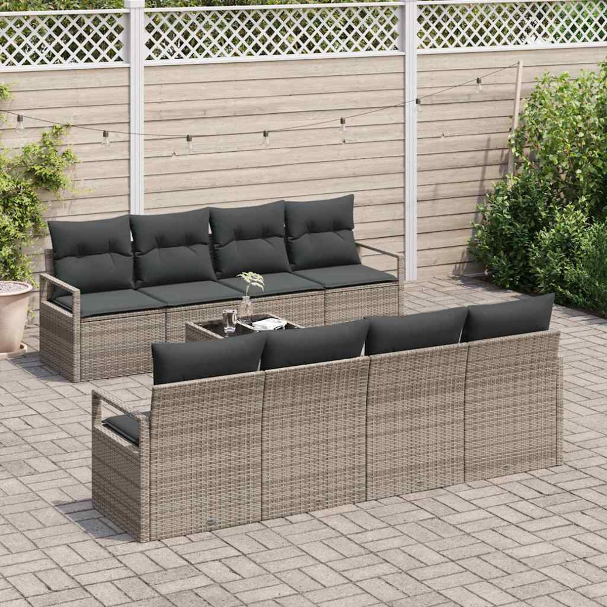 Escena de patio con muebles de ratán. Dos sofás con cojines grises, una mesa y un patio de ladrillo. Valla de madera al fondo.
