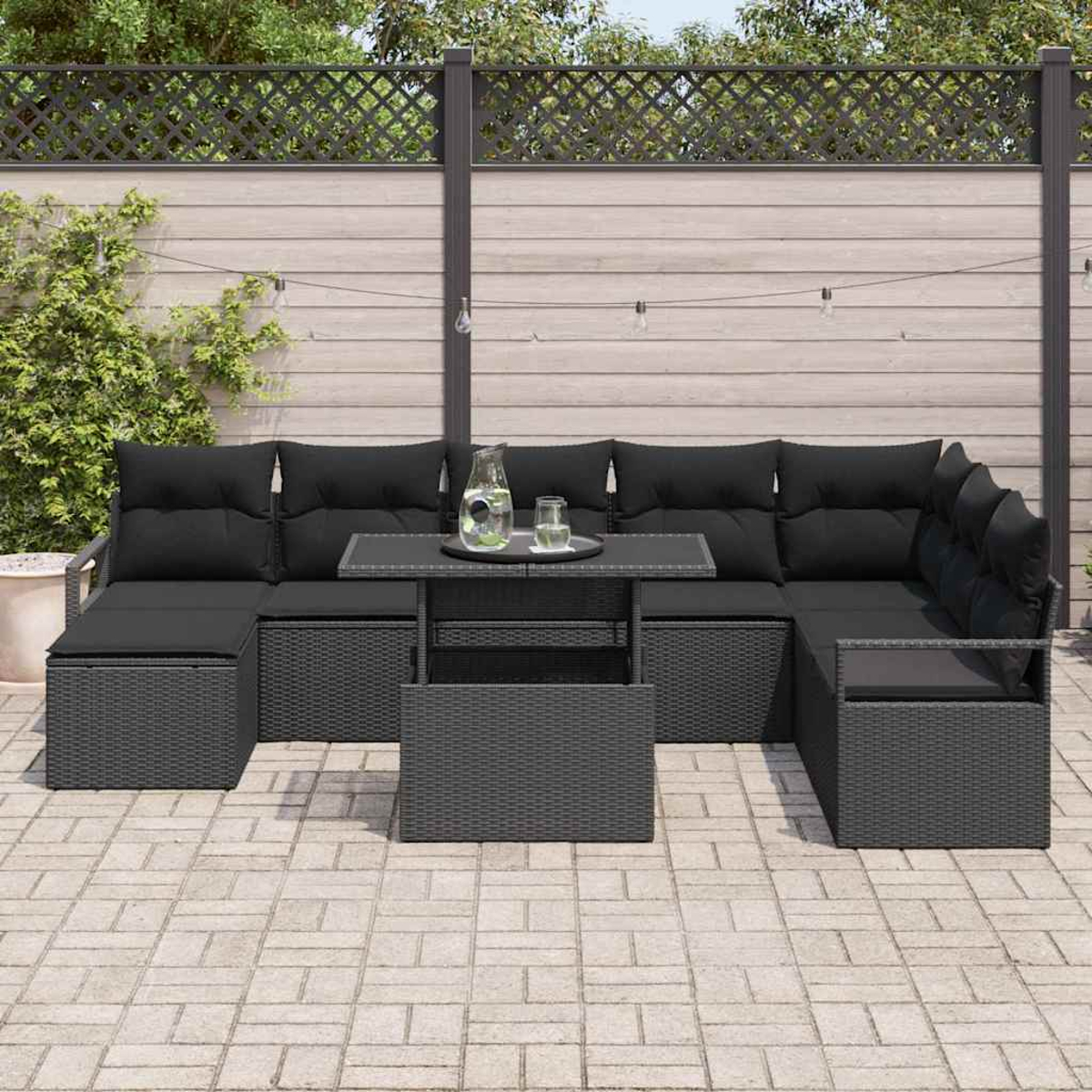 Garten-Terrassenset mit schwarzen Kissen und einem Tisch. Das Set ist grau, auf einer Backsteinterrasse. Im Hintergrund ein Zaun und Grün.