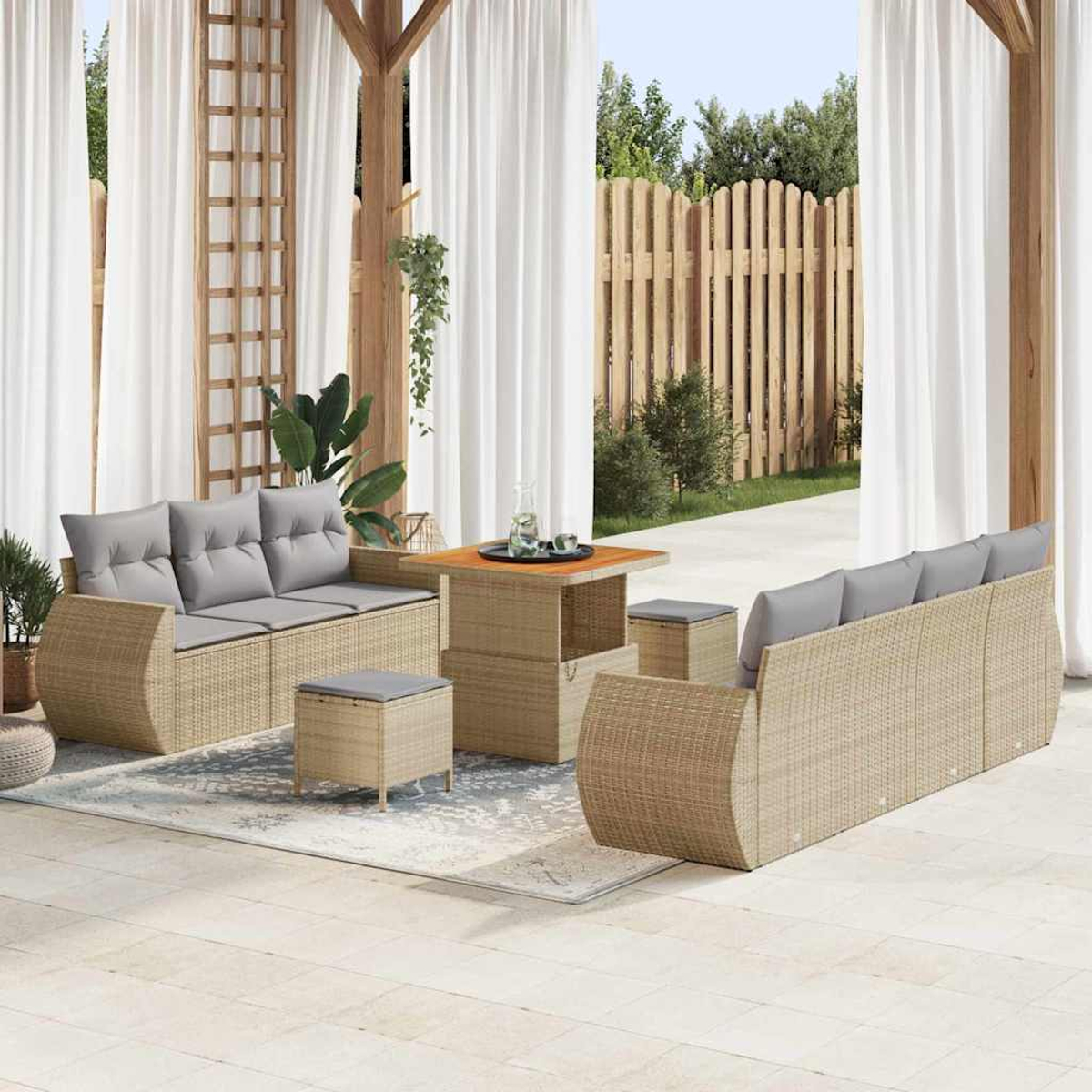 Garten-Terrassenset mit Korbs Sofas, grauen Kissen, Tisch und Hockern auf einem gemusterten Teppich. Weiße Vorhänge und ein Holzzaun sind sichtbar.