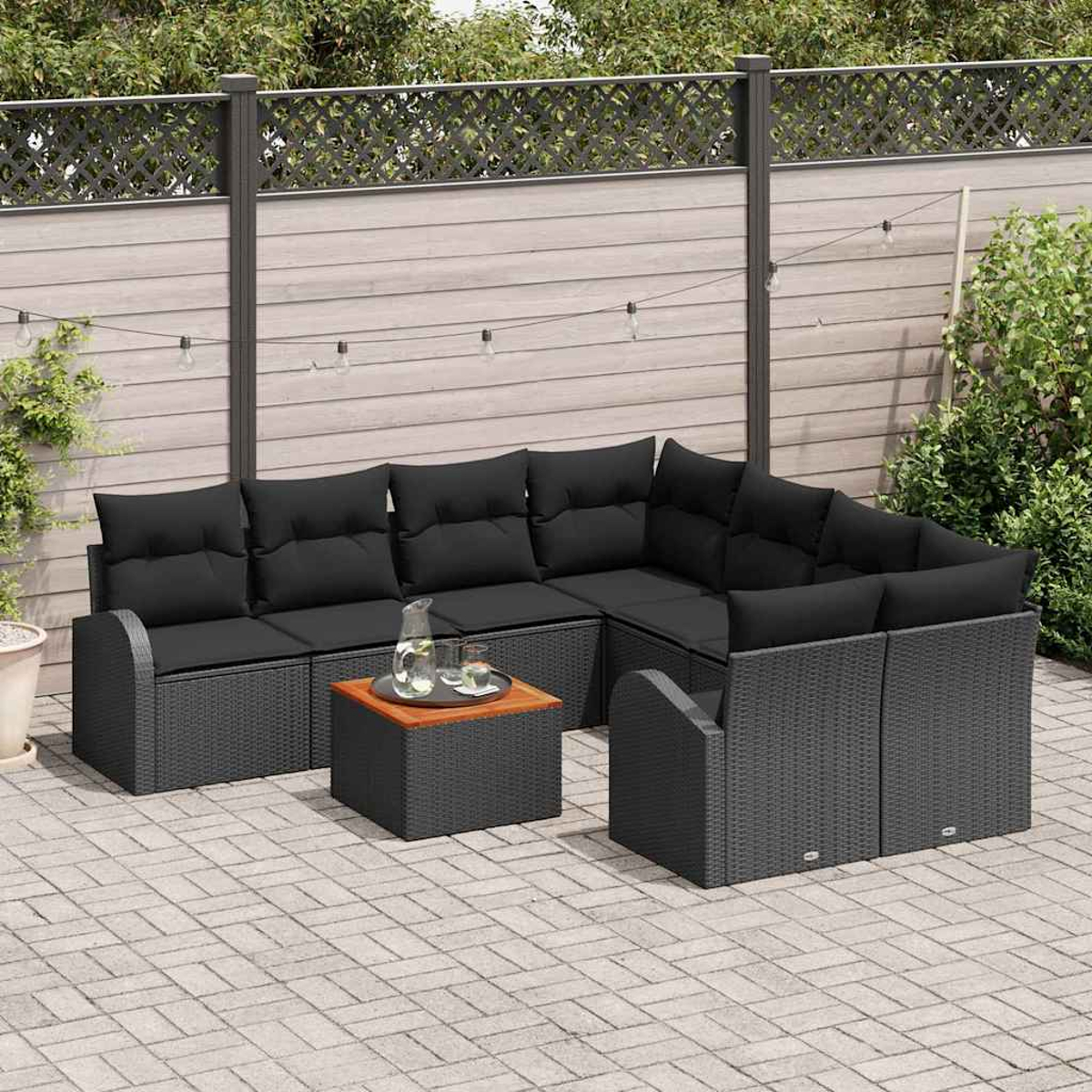 Garten-Sitzgruppe mit schwarzen Kissen auf grauem Korbgewebe, einem kleinen Tisch und einer Holzplatte. Im Hintergrund ein Zaun und Grün.