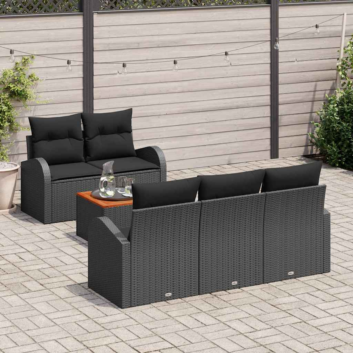 Terrassenmöbel-Set mit grauem Korbmöbel, schwarzen Kissen und Holztisch. Hintergrund mit Zaun und Lichterketten.