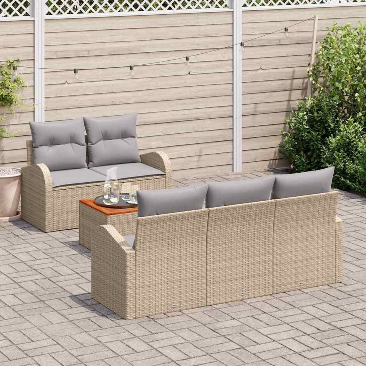 Terrassen-Set: beiges Korbmöbel, Zweisitzer mit grauen Kissen und Holztisch. Holzzaun und Grün im Hintergrund.