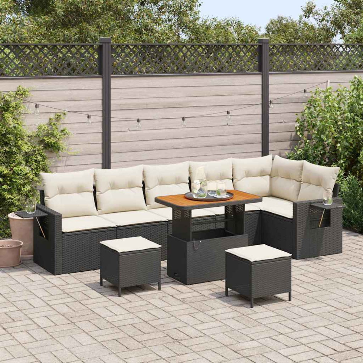 Garten-Terrassenset mit dunkelgrauem, gewebtem Sofa, cremefarbenen Kissen, Holztisch und zwei Hockern. Auf einer gepflasterten Terrasse mit Holzzaun.