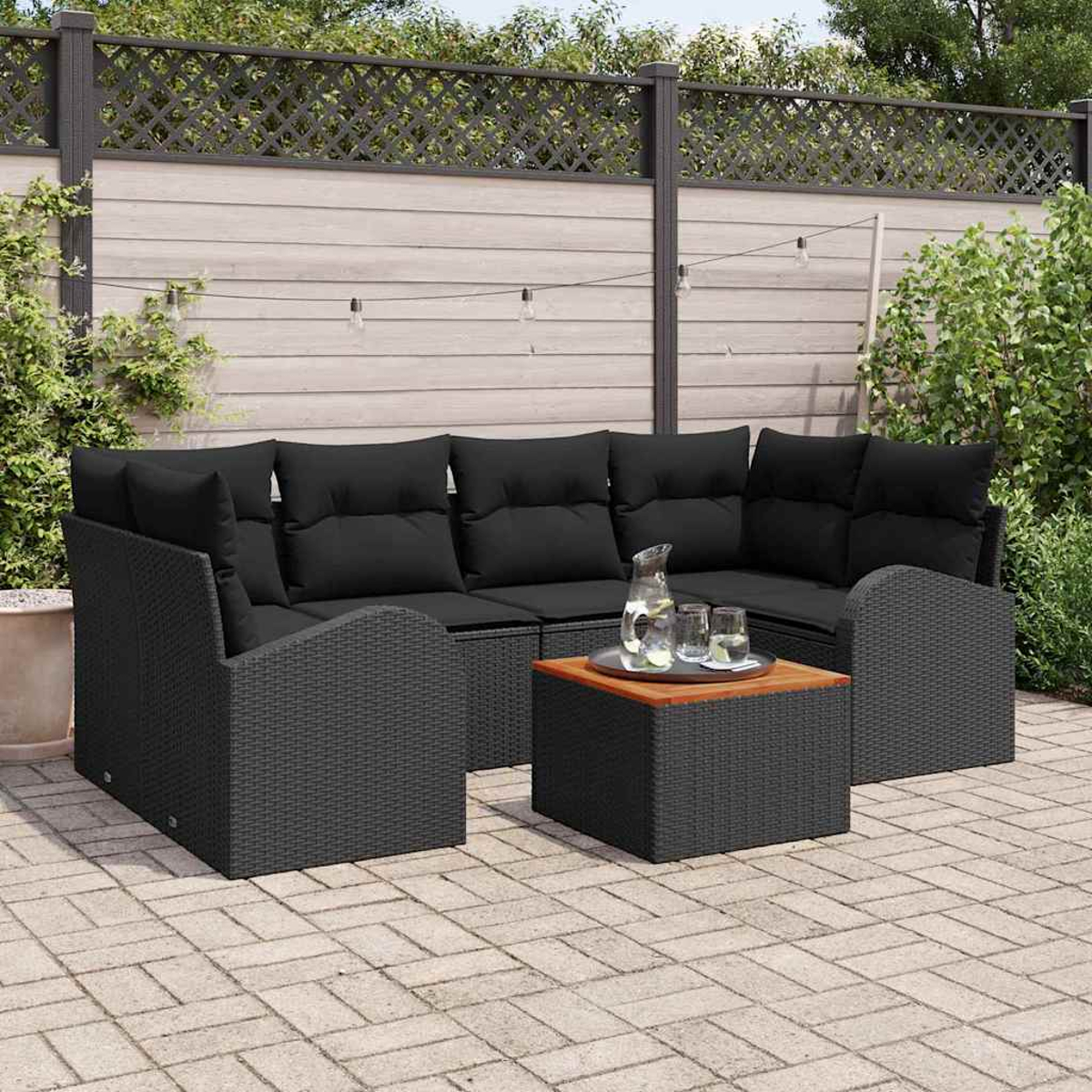 Outdoor-Sitzgruppe mit schwarzen Kissen, Tisch und Krug mit Gläsern. Auf einer Ziegelterrasse mit Zaun im Hintergrund.