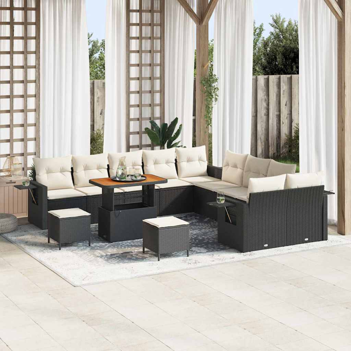 Zona de estar al aire libre con muebles de mimbre, cojines beige, mesa de madera y cortinas blancas. Incluye alfombra y vegetación, bajo estructura de madera.