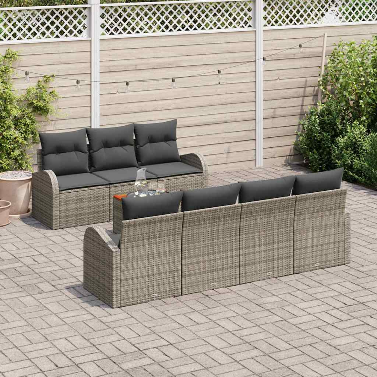 Szene im Freien mit grauen Korbmöbeln. Zwei Sofas mit dunkelgrauen Kissen stehen auf einer Ziegelpflasterung, im Hintergrund ein weißer Zaun.