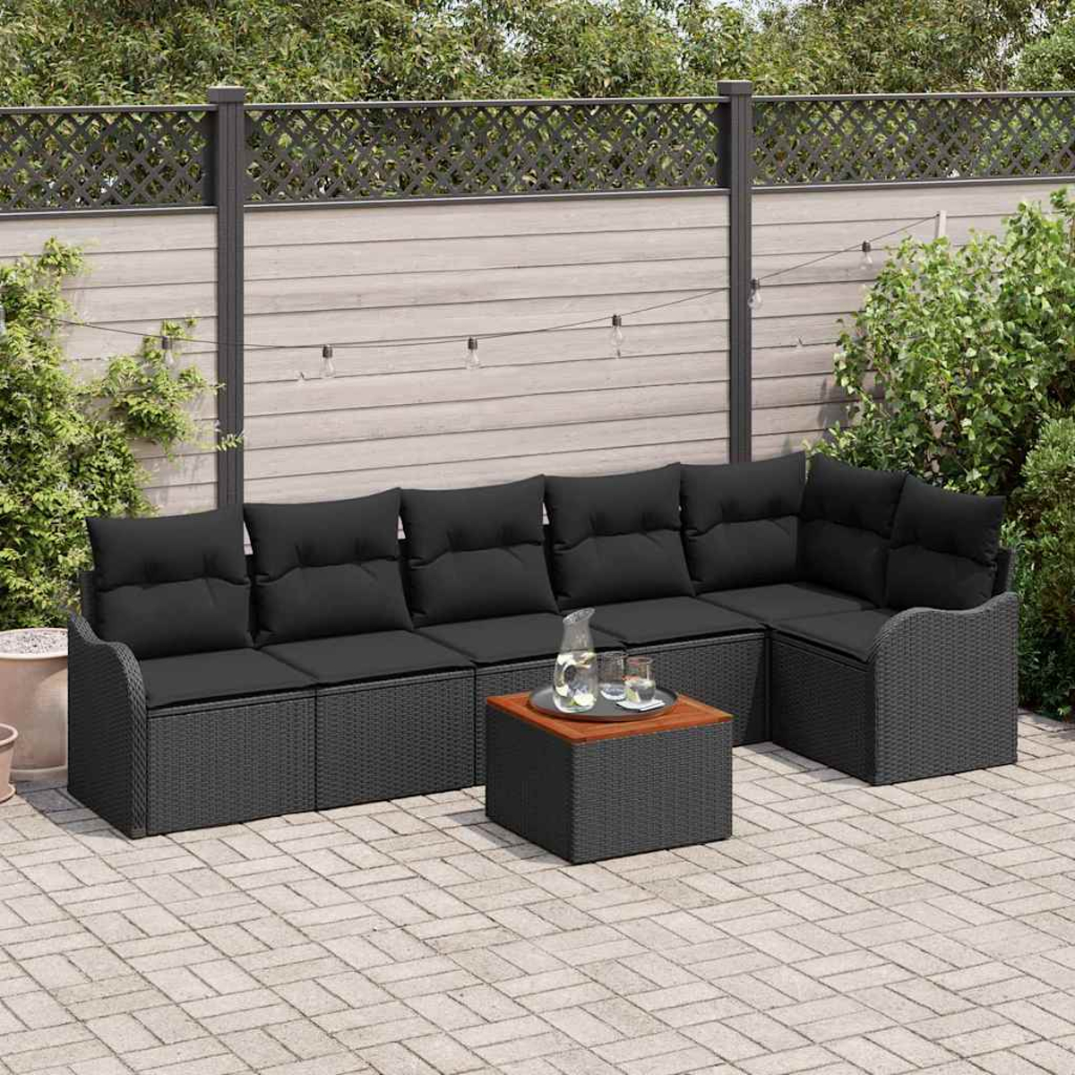 Schwarzes Korbsessel-Sofa mit Kissen und passendem Tisch stehen auf einer Ziegelterrasse. Hintergrund ist ein Zaun und Grün.