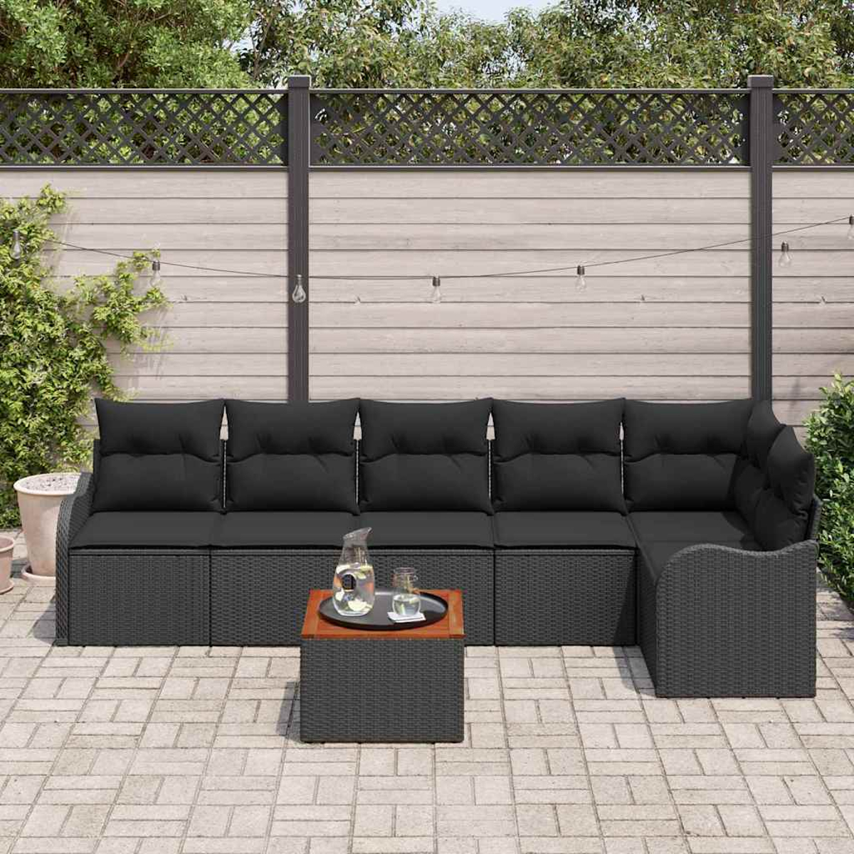 Ein Outdoor-Sofa aus schwarzem Korbgeflecht mit schwarzen Kissen und einem kleinen Tisch mit Getränken. Ein Holzzaun und Bäume sind im Hintergrund zu sehen.