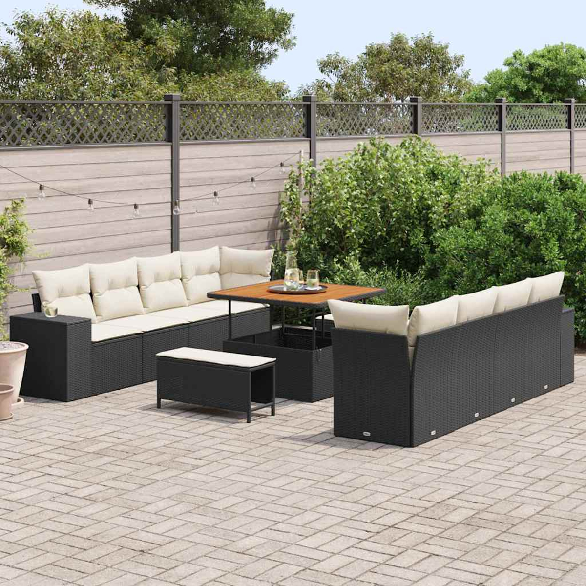 Garten-Set: schwarze Korbsessel, Tisch und Bank mit cremefarbenen Kissen, auf einer Terrasse, mit Zaun und Grün im Hintergrund.