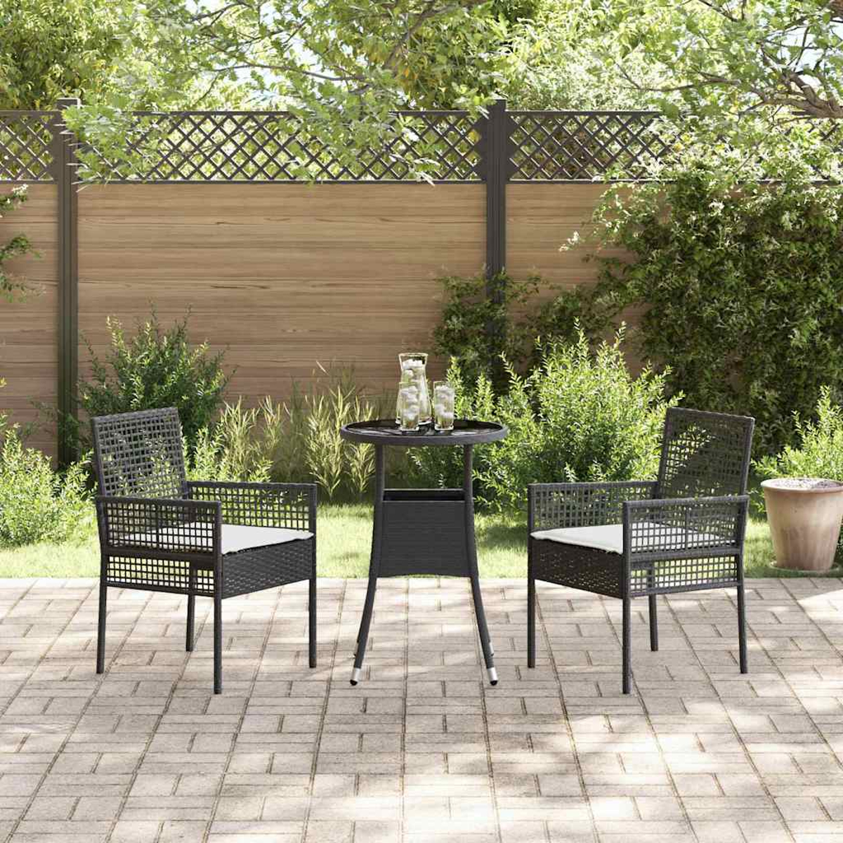 Eine Szene auf der Terrasse mit zwei Stühlen, einem Tisch und einem Zaun. Auf dem Tisch stehen Gläser und eine Kanne. Im Hintergrund grünes Laub.