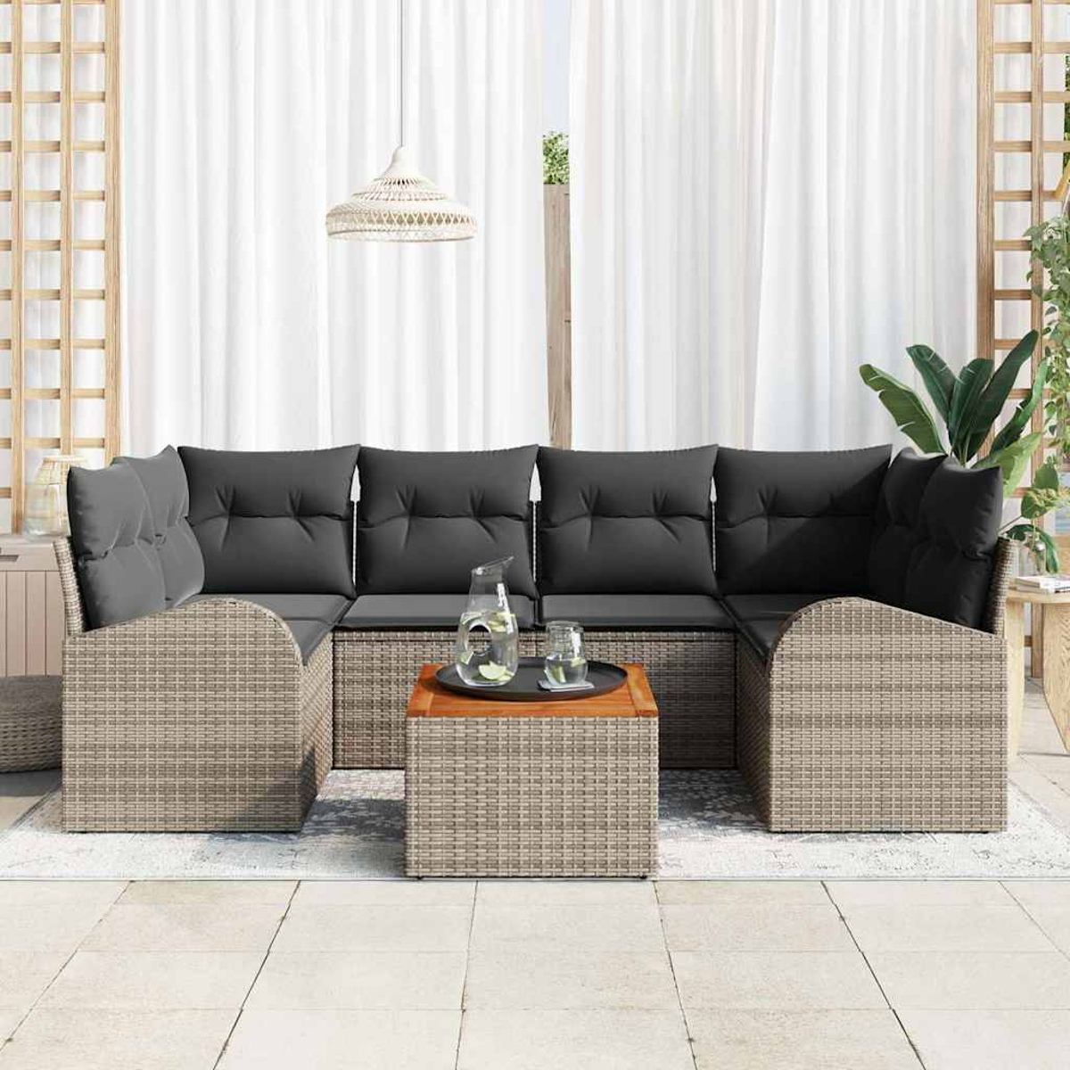 Ein Rattansofa mit grauen Kissen, ein Holztisch mit Wasser und Gläsern darauf und weiße Vorhänge im Hintergrund.