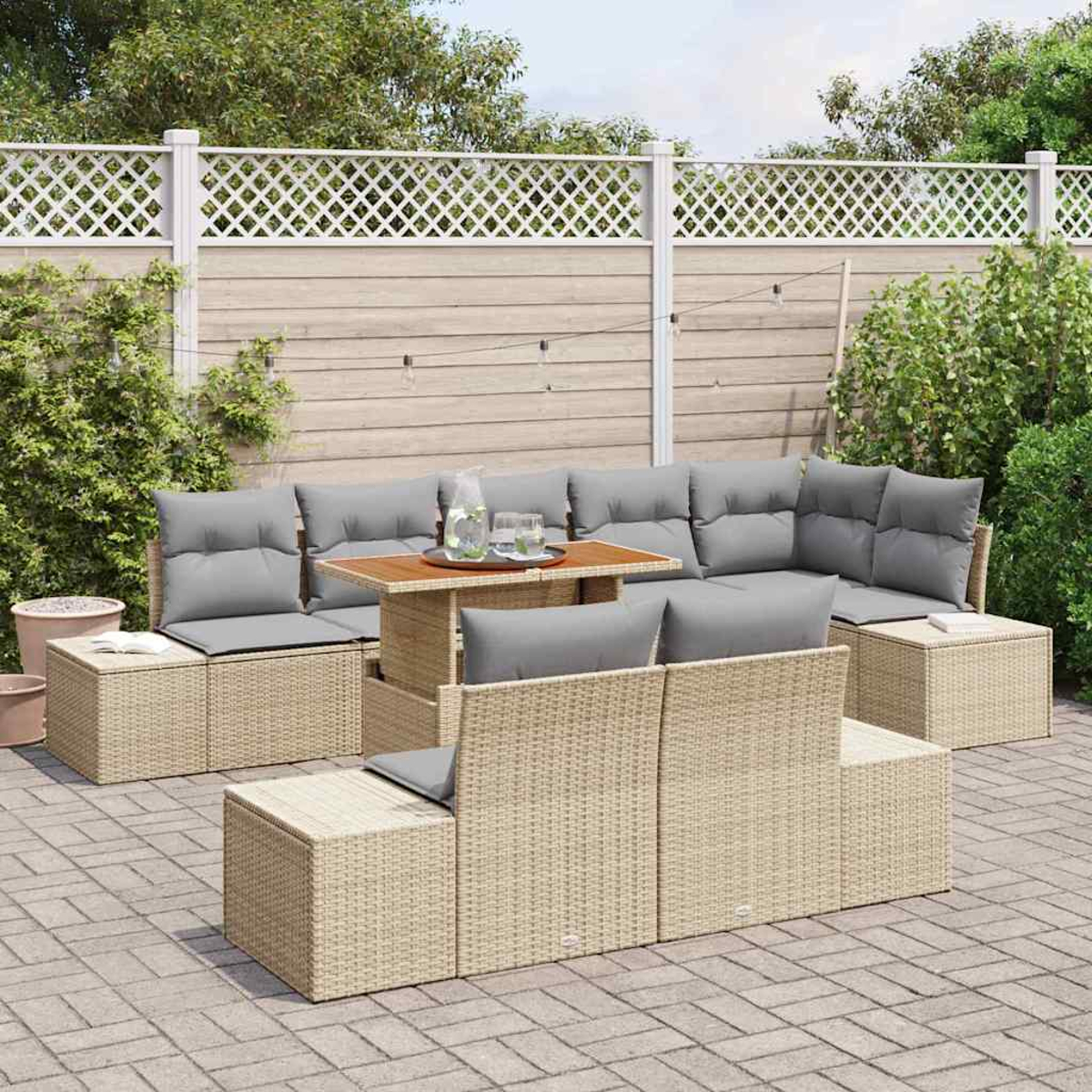 Gartenmöbel-Set aus Korbgeflecht mit grauen Kissen und einem Holztisch auf einer gepflasterten Terrasse, mit weißem Gitterzaun im Hintergrund.