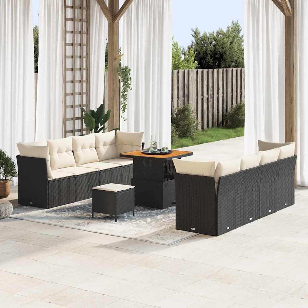 Outdoor-Sitzbereich mit schwarzem Korbmöbel, cremefarbenen Kissen, einem quadratischen Tisch und durchsichtigen weißen Vorhängen. Im Hintergrund ein Zaun.