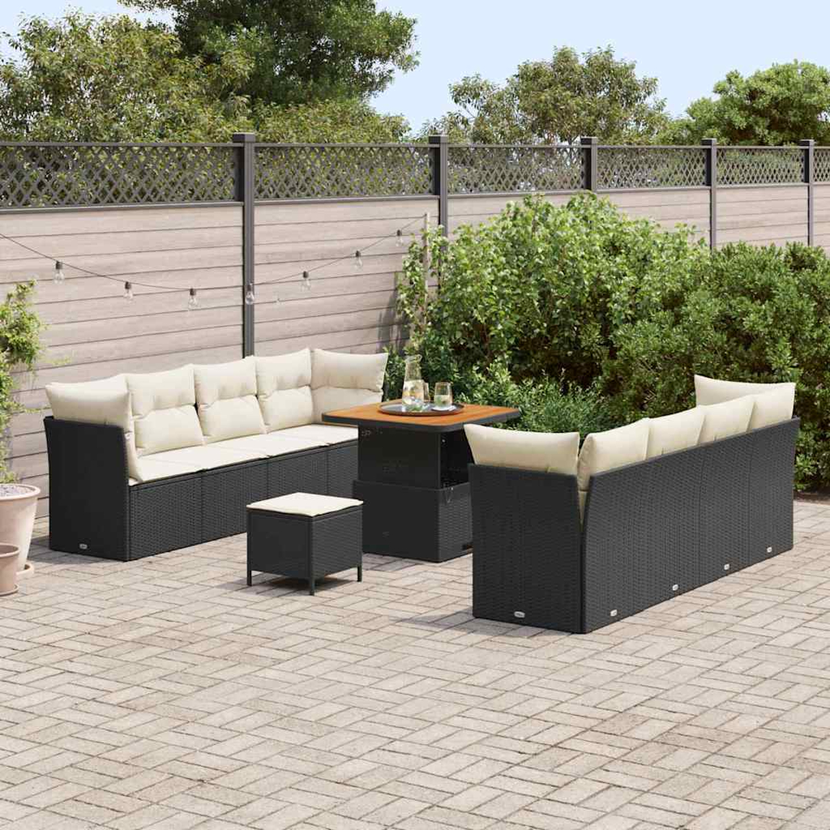 Garten-Terrassenset mit schwarzen Korbmöbeln, beigen Kissen und einem Holztisch. Vor einem Holzzaun und grünen Büschen.