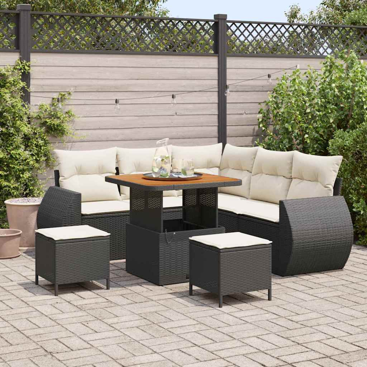 Terrassenset im Freien. Dunkelgraues Korbmöbel-Sofa, Tisch und Hocker mit cremefarbenen Kissen. Holzzaun und grüne Pflanzen im Hintergrund.