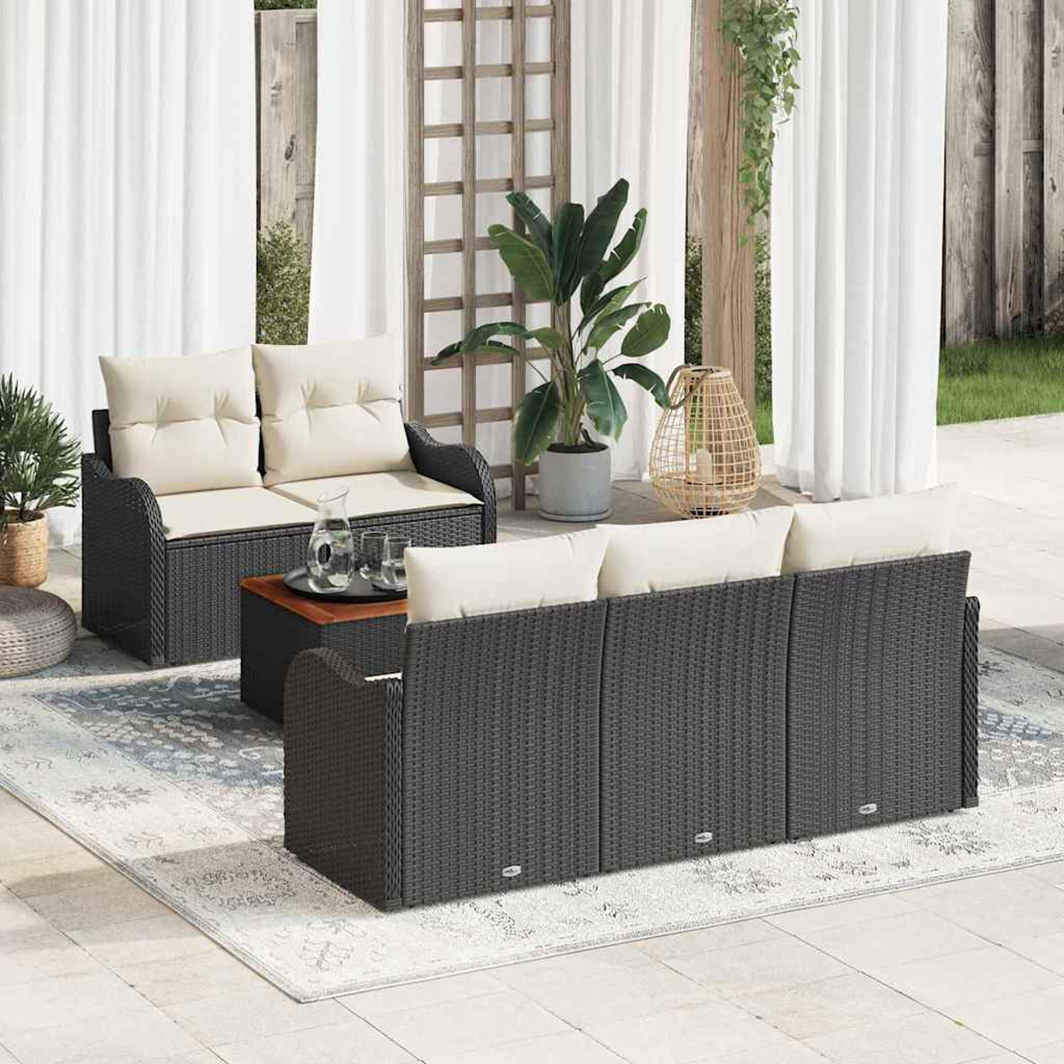 Szene im Freien mit schwarzen Korbmöbeln, cremefarbenen Kissen und einem Holztisch. Große Pflanzen, weiße Vorhänge und ein Teppich.