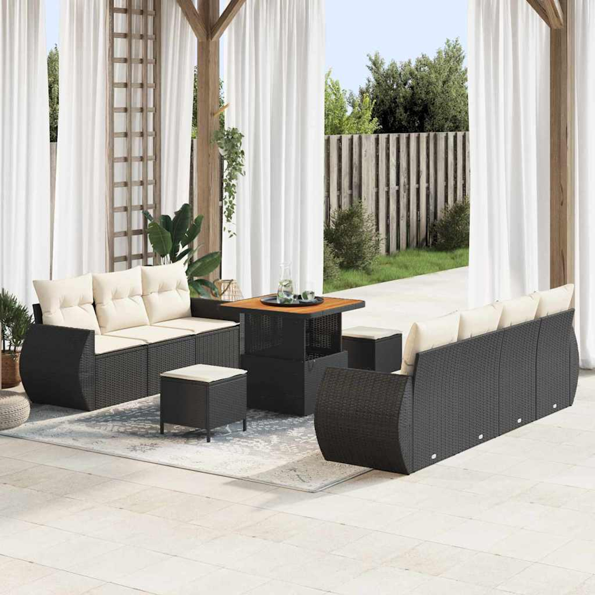 Juego de muebles de patio exterior con sofás de mimbre negro, cojines beige, una mesa cuadrada y cortinas blancas al fondo.