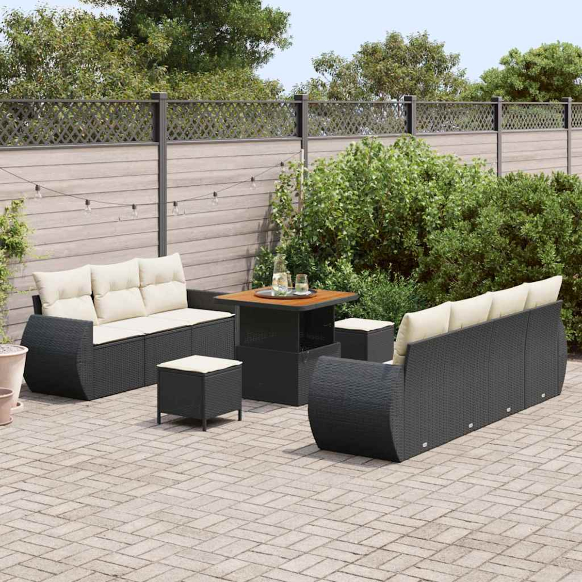 Juego de muebles de patio: sofás de mimbre negro, otomanas y mesa con cojines beige. Sobre patio de ladrillos, con vegetación y valla.