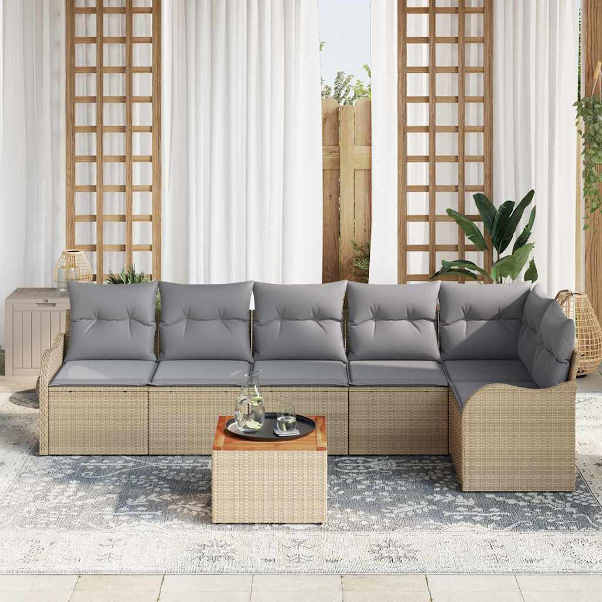 Outdoor-Ecksofa aus Korbgeflecht mit grauen Kissen, kleiner Tisch mit Getränken, auf gemustertem Teppich. Dahinter weiße Vorhänge und Holzgitter.