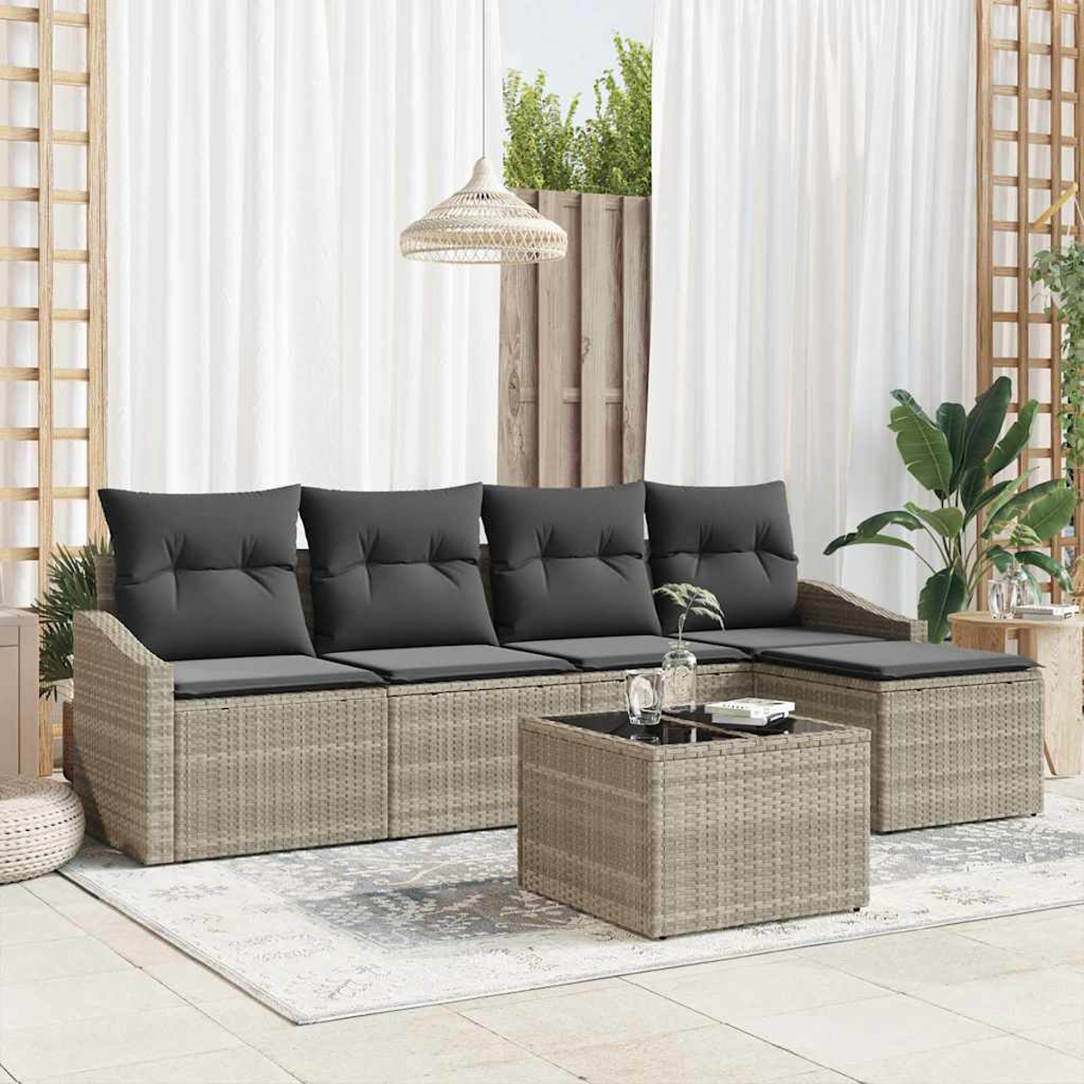 Outdoor-Korbmöbel mit grauen Kissen, einem passenden Couchtisch und einem gemusterten Teppich auf einer Terrasse. Vorhänge und Pflanzen im Hintergrund.