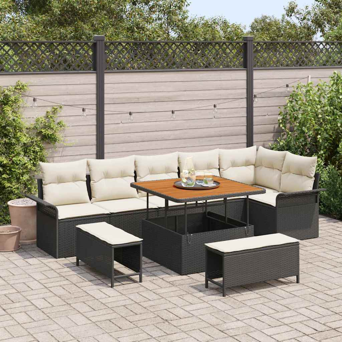 Garten-Terrassenset mit schwarzem Korbmöbel, beigen Kissen und Holztisch. Im Hintergrund ein Zaun und Grün.