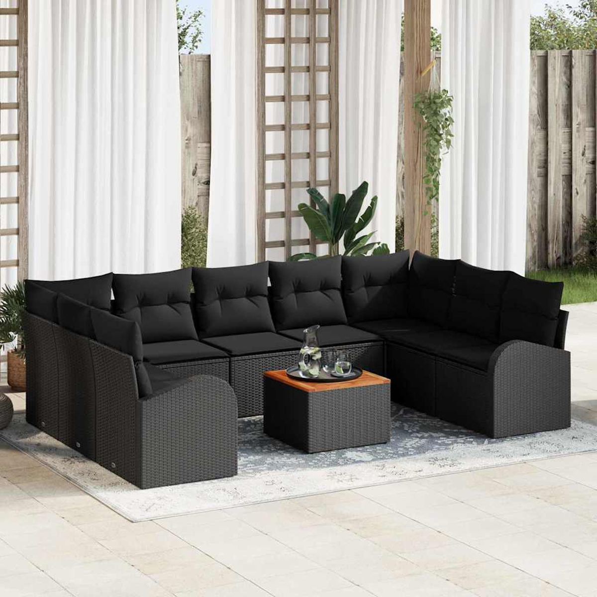 Garten-Set mit schwarzen Korbmöbeln, Kissen und einem Couchtisch mit Holzplatte auf einem Teppich. Weiße Vorhänge im Hintergrund.