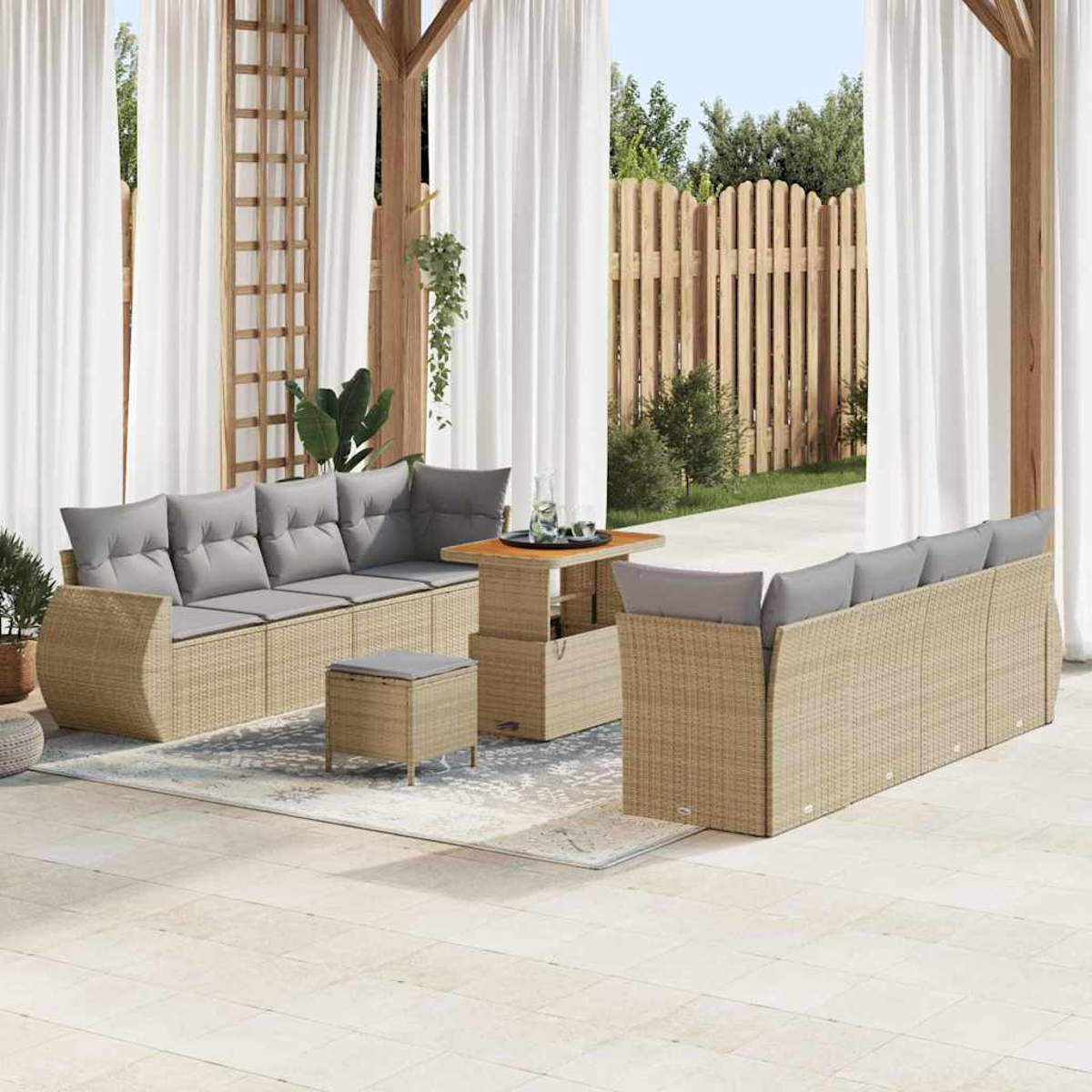 Gartenmöbel-Set aus Korbgeflecht. Graue Kissen auf Sofas, Ottomanen und Tisch. Weiße Vorhänge, Holzpergola und Zaun im Hintergrund.