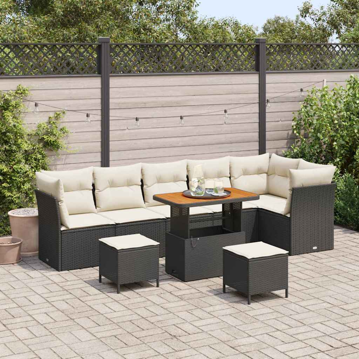 Garten-Terrassenset mit Sofa, zwei Ottomanen und einem Tisch. Beige Kissen kontrastieren mit dem dunkelgrauen Korbgeflecht. Tischplatte aus Holz und Zaun.