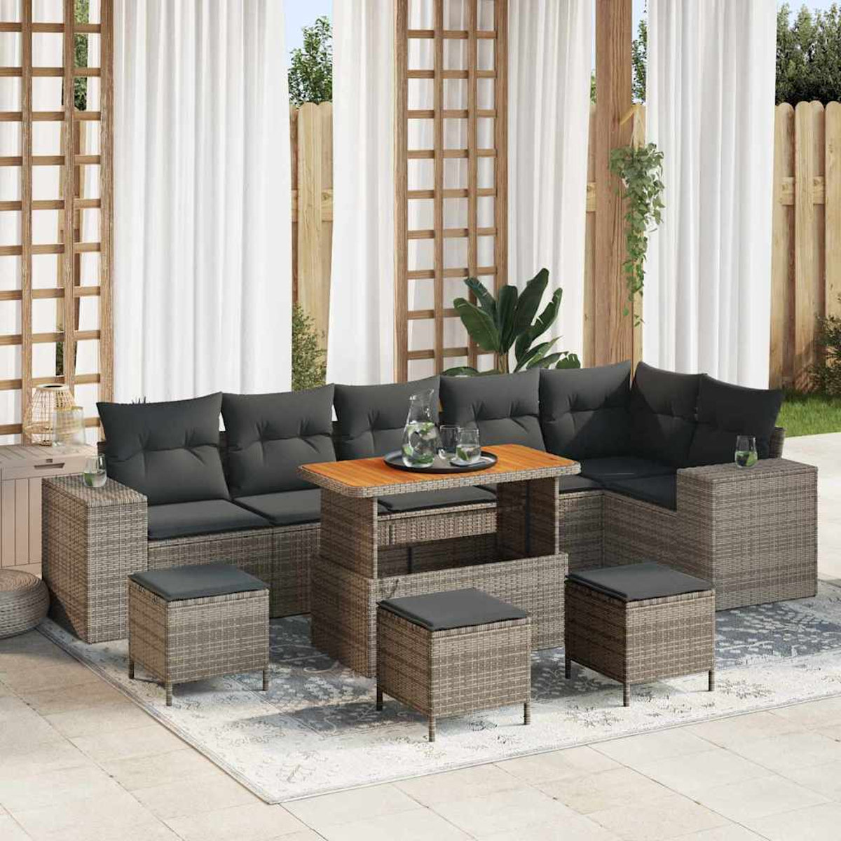 Set di mobili da esterno: divano angolare grigio con cuscini neri, tavolo in legno e pouf su un tappeto, con tende bianche sullo sfondo.