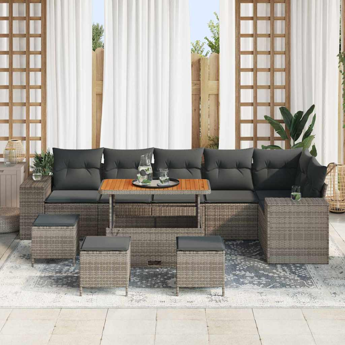 Set da patio esterno con cuscini grigi, tavolo in legno e mobili intrecciati grigi. Tende bianche e una staccionata in legno sullo sfondo.
