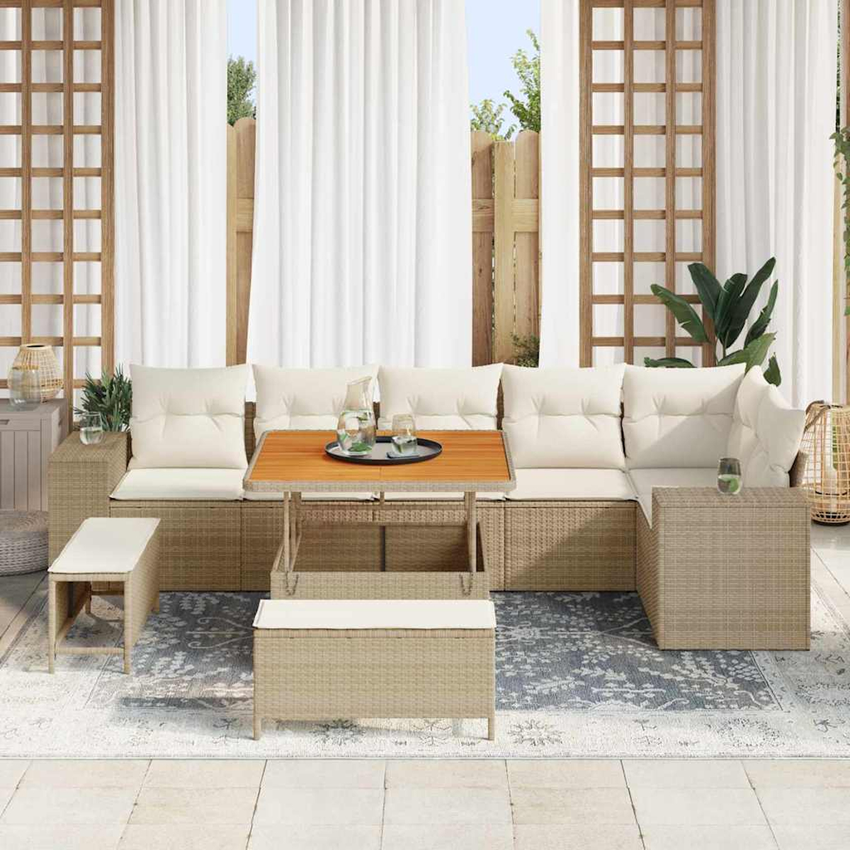 Area salotto all'aperto con mobili in rattan beige, cuscini bianchi, tavolo in legno e tende bianche sullo sfondo.
