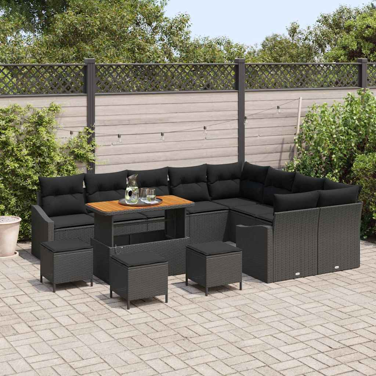 Garten-Terrassenset mit schwarzen Kissen. Enthält Ecksofa, Hocker und einen Holztisch mit Getränken. Vor einem Zaun und Grün.