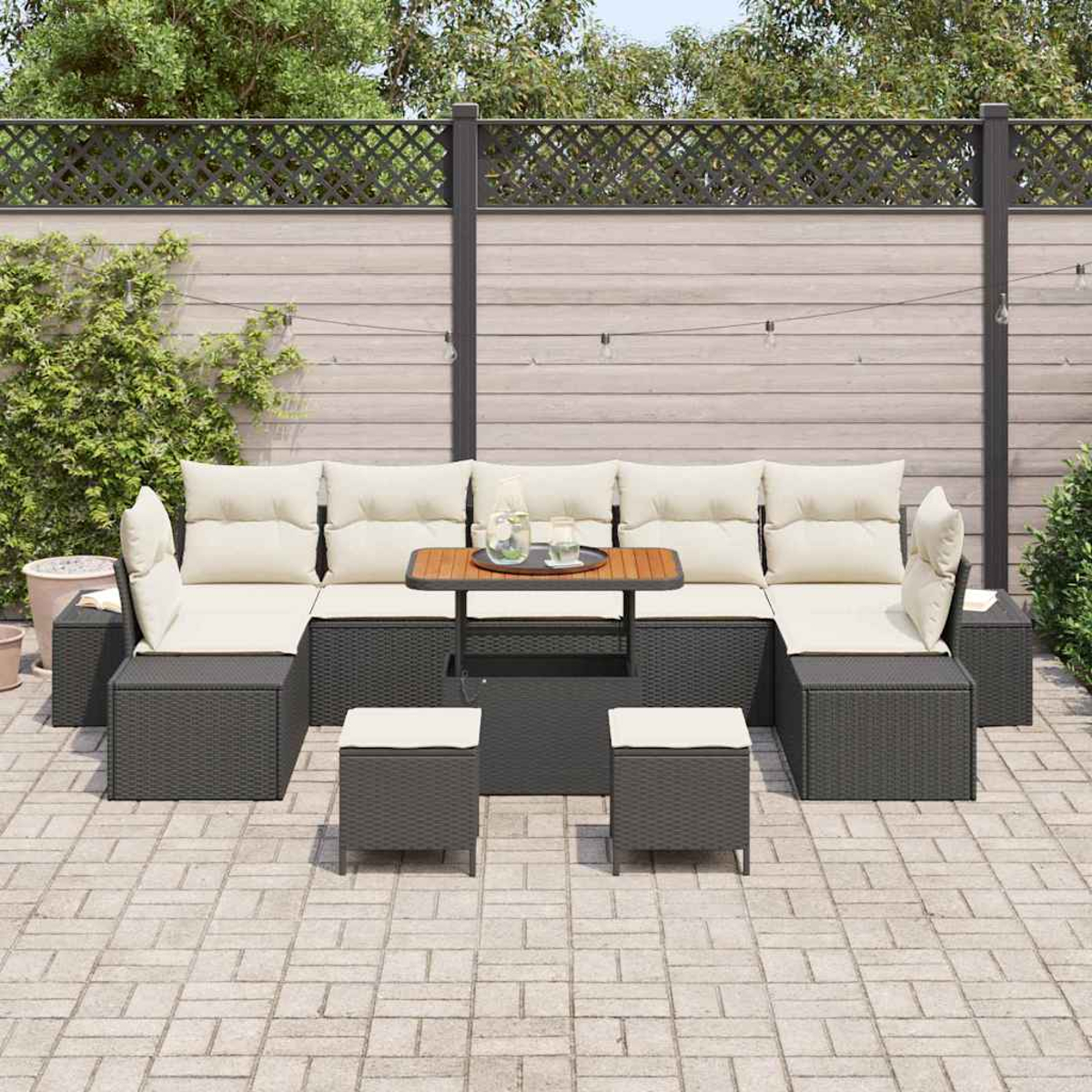 Ein Outdoor-Terrassenset mit dunklem Korbmöbel, cremefarbenen Kissen, einem kleinen Tisch und zwei Ottomanen auf einer Backstein-Terrasse, mit Zaun im Hintergrund.