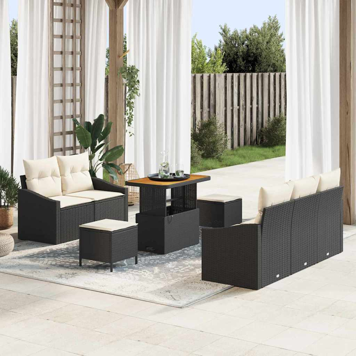 Terrassenmöbel-Set mit schwarzen Korbmöbeln, cremefarbenen Kissen, Couchtisch und Teppich. Weiße Vorhänge und Holzzaun im Hintergrund.