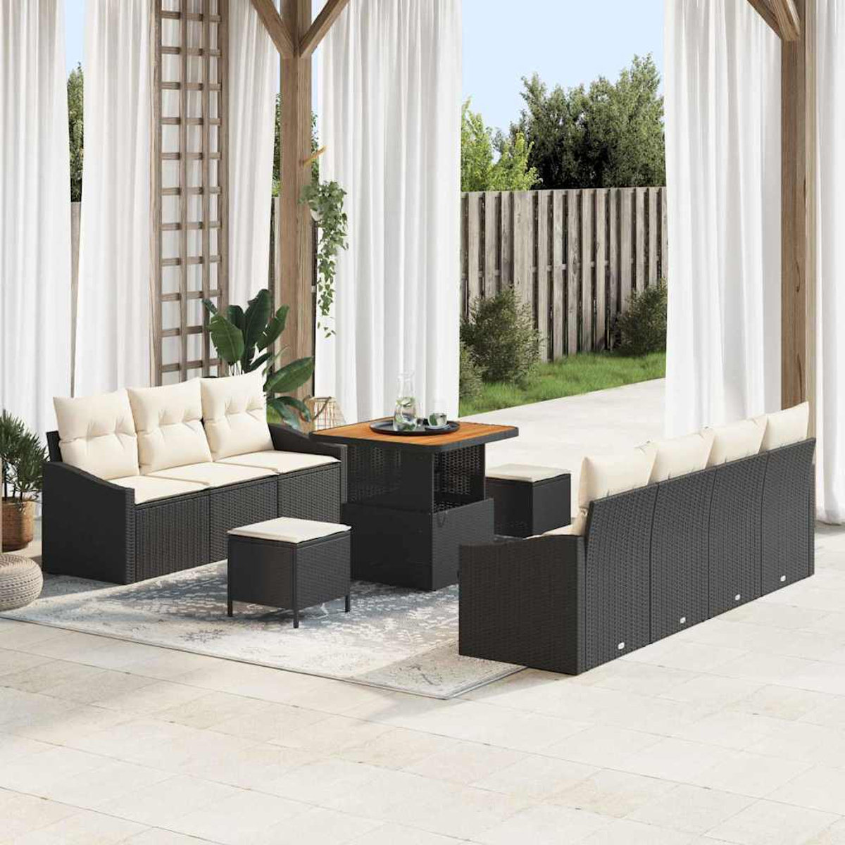 Terrassenmöbel-Set im Freien mit schwarzen Korbmöbeln, beigen Kissen, Holztisch und weißen Vorhängen. Im Hintergrund grüne Pflanzen und ein Holzzaun.