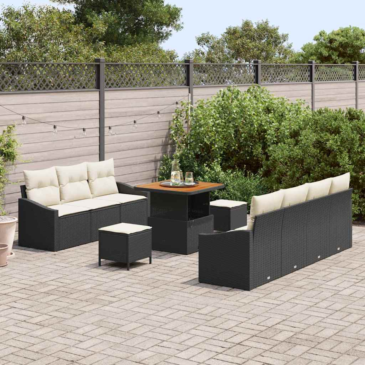 Terrassenmöbel-Set. Schwarze Korbmöbel mit cremefarbenen Kissen. Enthält Sofa, zwei Stühle, einen Tisch und zwei Hocker. Grüner Hintergrund.