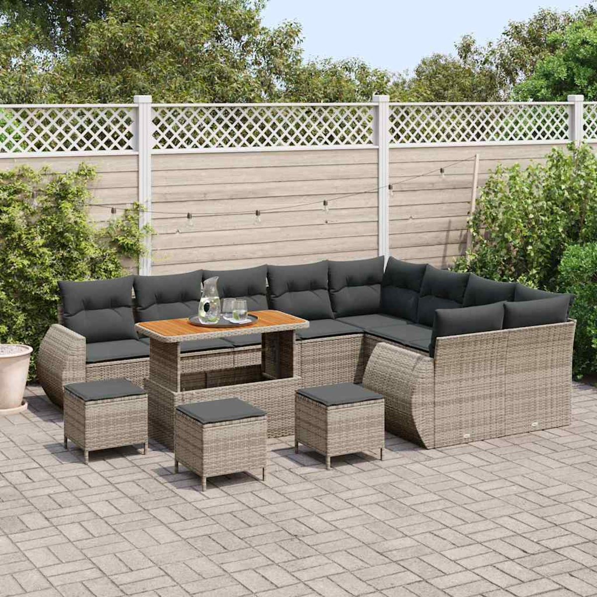 Outdoor-Wicker-Ecksofa mit Ottomanen, einem Holztisch und Kissen. Auf einer gepflasterten Terrasse mit weißem Zaun und Grün.