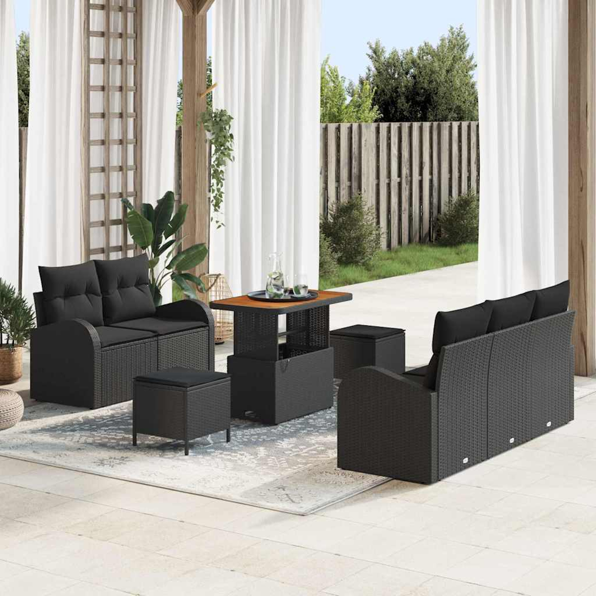 Terrassenset im Freien mit schwarzen Korbsesseln, Ottomanen und einem Tisch. Weiße Vorhänge und ein Holzzaun im Hintergrund.