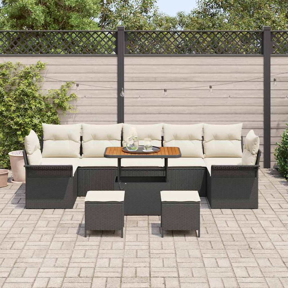 Terrassenset mit grauen Korbmöbeln, beigen Kissen und einem Holztisch. Auf einer Ziegelterrasse. Hintergrund umfaßt Zaun und Pflanzen.