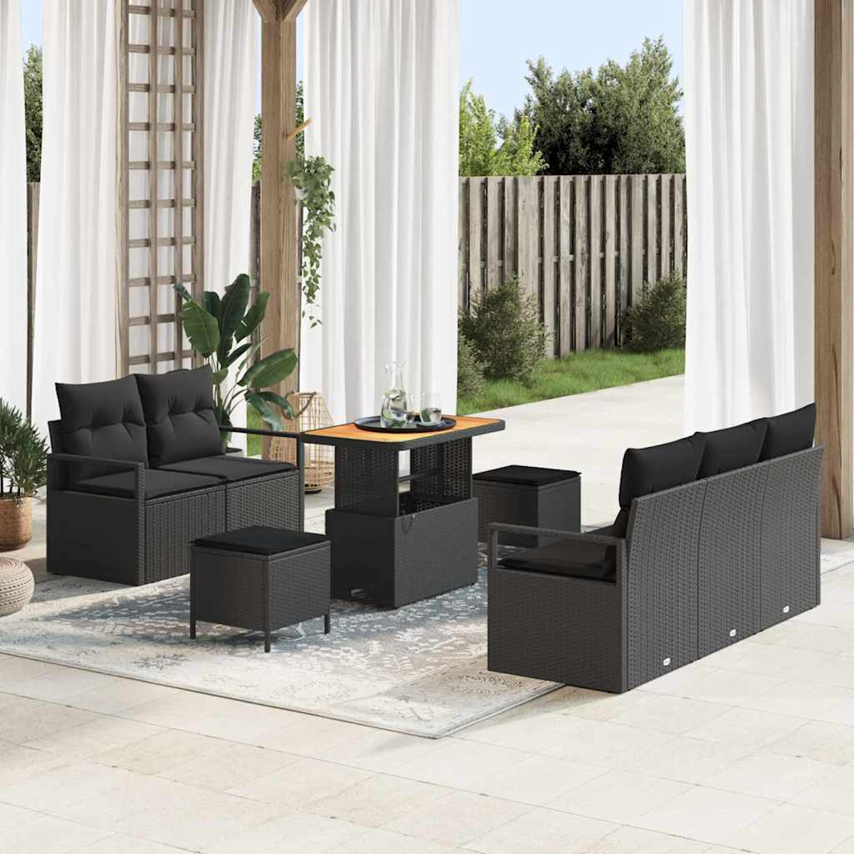 Terrassengarnitur mit schwarzem Korbmöbel, einem Tisch und Kissen. Weiße Vorhänge und ein Holzzaun im Hintergrund.