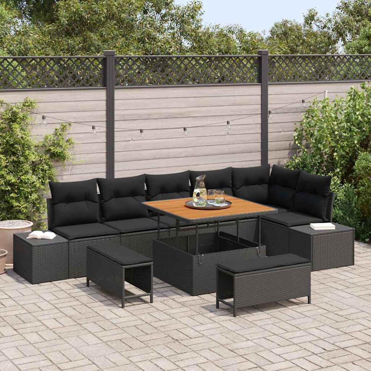 Juego de patio al aire libre con cojines negros y una mesa de madera. Rodeado de una valla, plantas y luces de cadena. Una jarra y vasos están en la mesa.