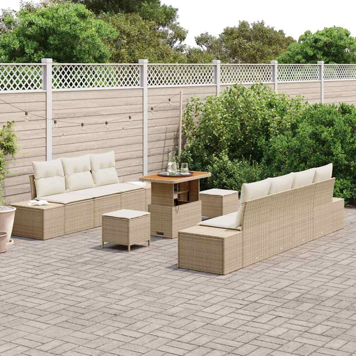 Garten-Terrassenset mit beigen Korbsesseln, Ottomanen und einem Tisch auf einer gepflasterten Terrasse. Ein weißer Zaun und üppiges Grün im Hintergrund.
