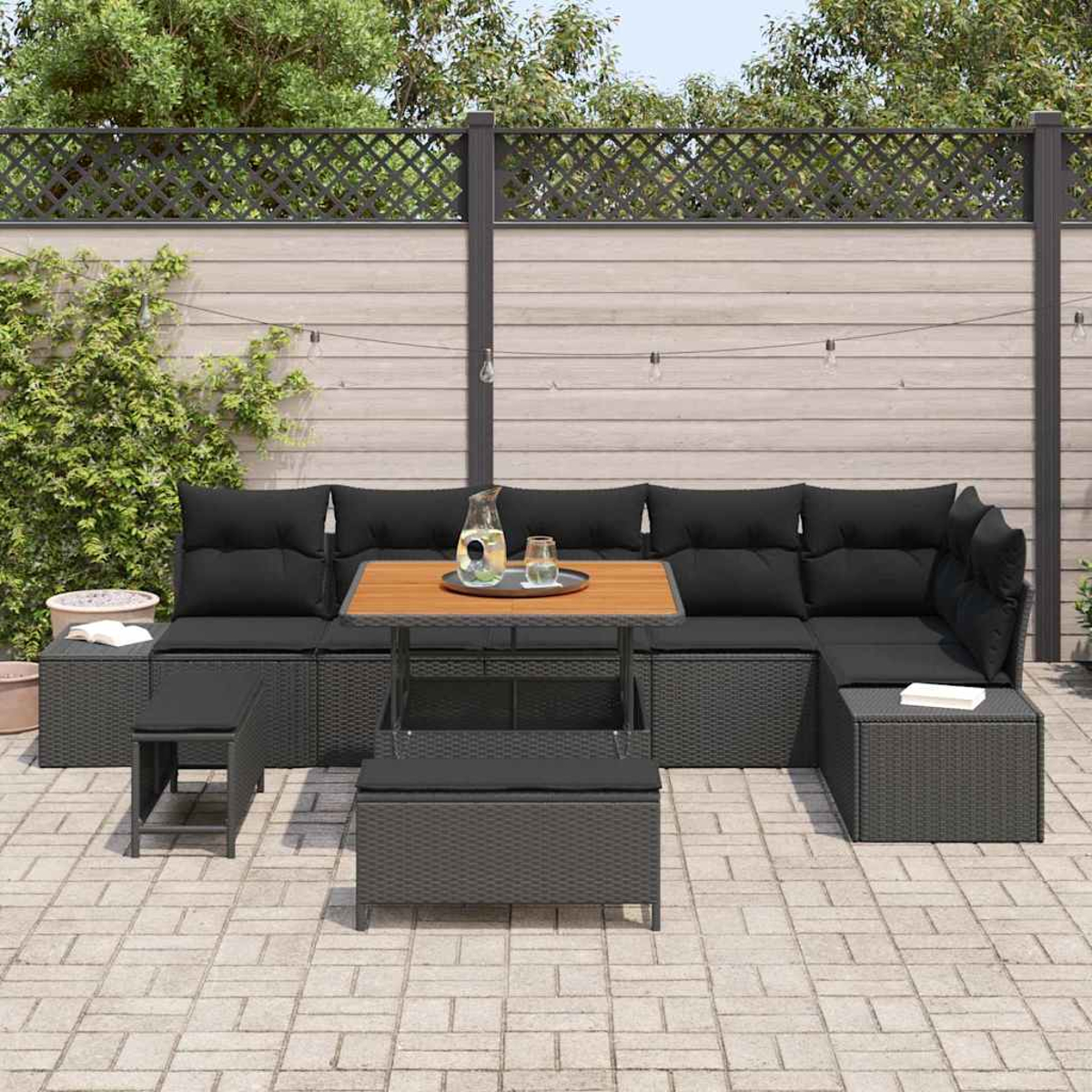 Sofá seccional exterior con cojines negros y mesa de madera. El fondo muestra una valla de madera y vegetación.