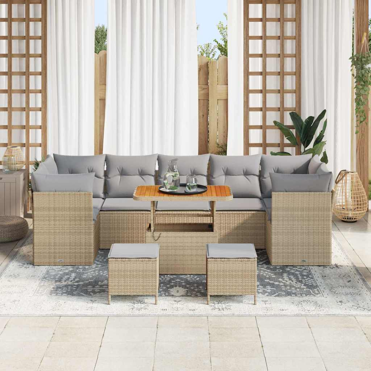 Divano in rattan da esterno con cuscini grigi, tavolo in legno e due pouf, posizionato su un tappeto fantasia e tende bianche sullo sfondo.