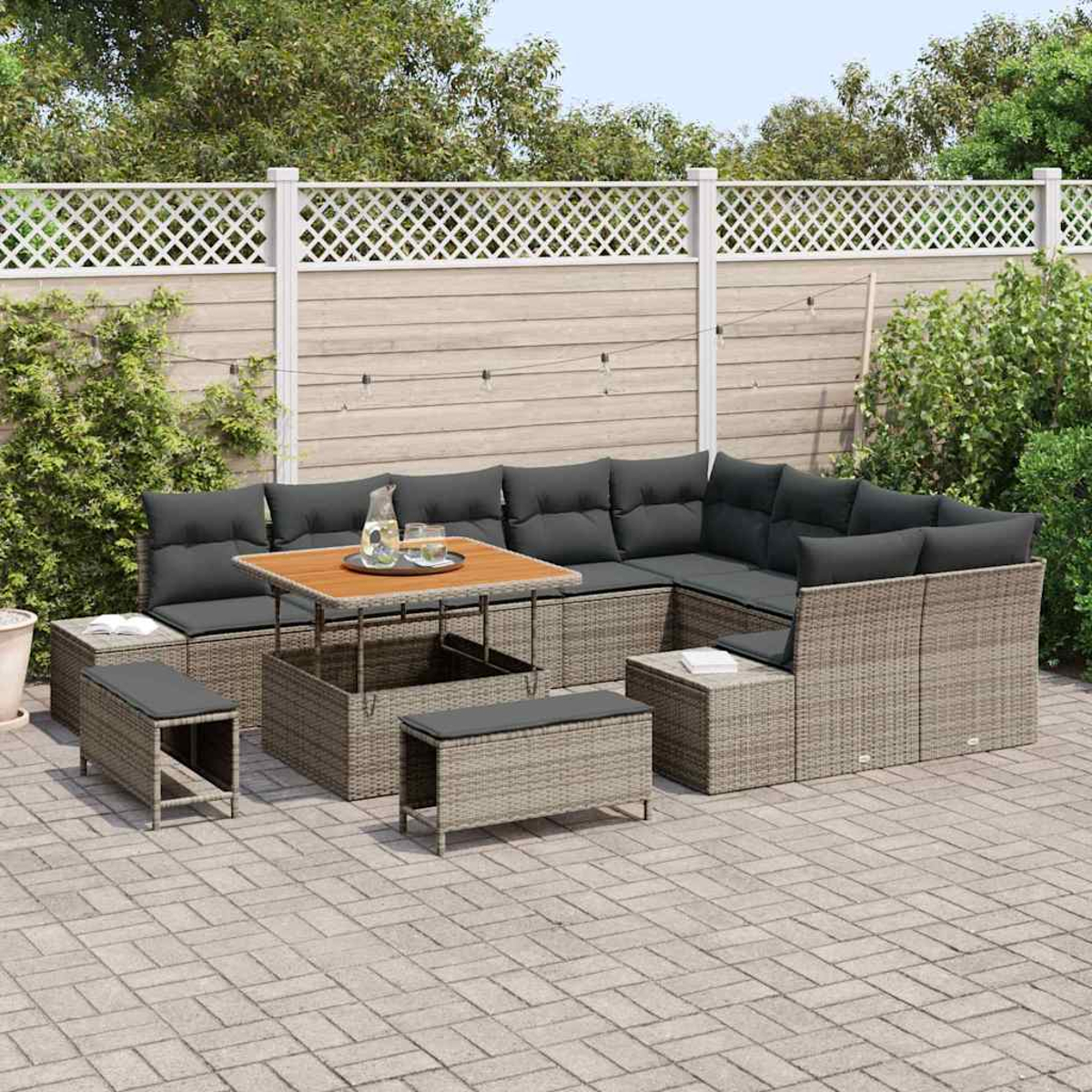 Ein Terrassenset mit grauen Kissen, einem Holztisch und Bänken auf einer Ziegelterrasse. Ein weißer Zaun und Grünzeug im Hintergrund.