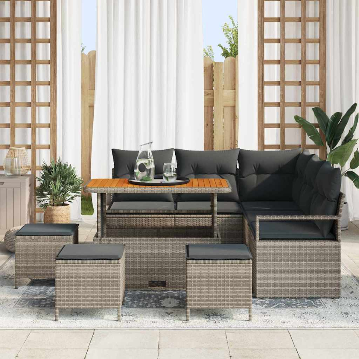 Terrassen-Set im Freien. Ein graues Korbmöbel-Sofa mit Kissen, ein Tisch mit Tablett und Hocker. Weiße Vorhänge und Pflanzen im Hintergrund.