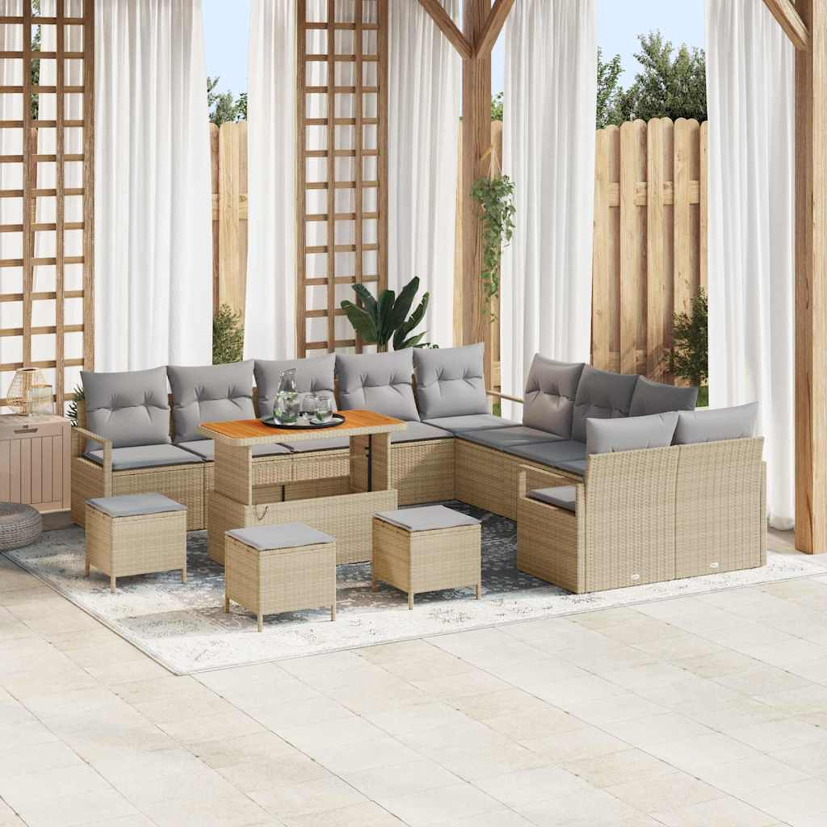 Juego de patio al aire libre con sofá, otomanas y mesa de centro. Cojines grises sobre muebles de mimbre beige. Cortinas blancas y estructura de madera.