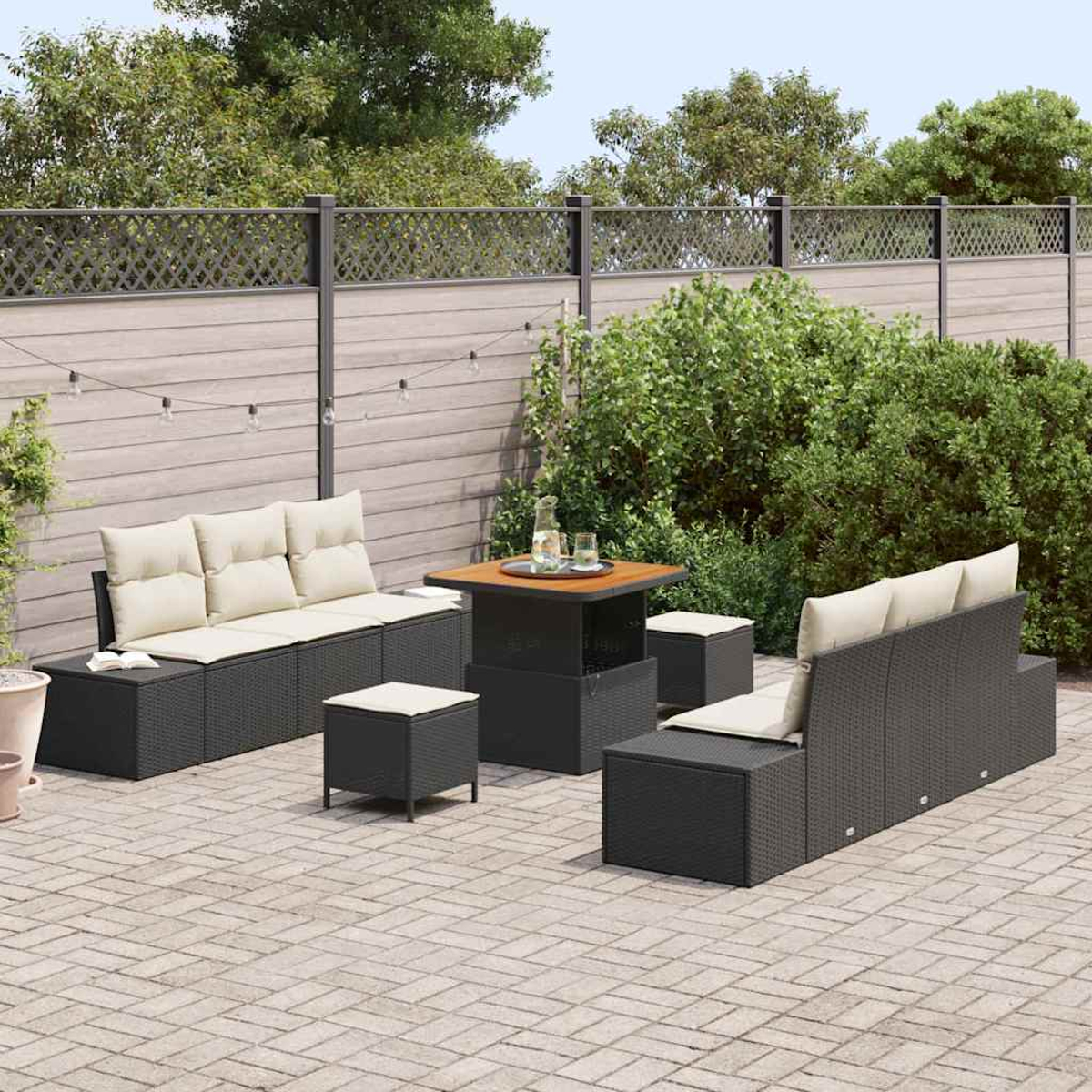 Garten-Terrassenset mit schwarzer Korbmöbel, beige Kissen. Tisch und Hocker. Grüne Sträucher und Bäume im Hintergrund. Ziegel-Terrasse.