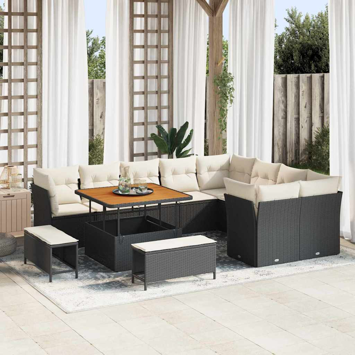 Terrassenset mit schwarzen Korbmöbeln, cremefarbenen Kissen, Holztisch und weißen Vorhängen. Bänke und ein Teppich vervollständigen das Set.
